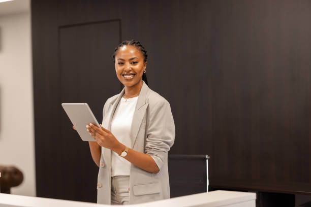 The young African-American female receptionist stands behind the reception desk with a digital tablet in her hands and smiles at the camera.